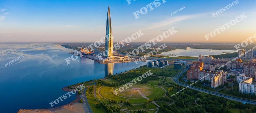 На фото изображен Лахта Центр — самый высокий небоскреб Европы, расположенный на берегу Финского залива в Санкт-Петербурге. Его стеклянный фасад отражает закатное небо, создавая эффектный визуальный акцент. На переднем плане зеленые зоны и жилые районы города, гармонично сочетающиеся с современным деловым кварталом. Финский залив добавляет простора и красоты к общей композиции.